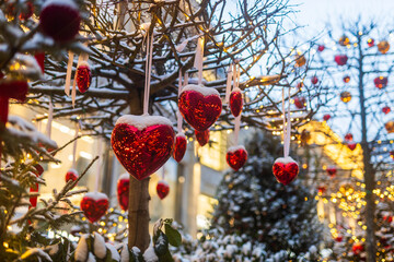 Close-up of a snow-covered Christmas tree decorated with red heart ornaments hanging on ribbons and warm lights.