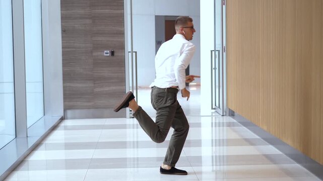 Happy young caucasian businessman in glasses and wireless earbuds dancing energetically in a modern office hallway, celebrating a successful deal, achievement, or promotion with funny moves