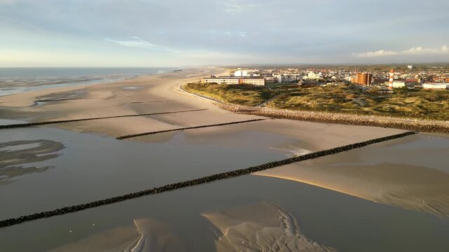 Drone aerial panorama of Berck-sur-Mer, France coastline with vast sandy beach at low tide, rock groynes and seawall, dunes and striped lighthouse glowing in warm evening light