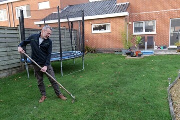 Man rakes autumn leaves on green lawn in backyard near brick house and trampoline