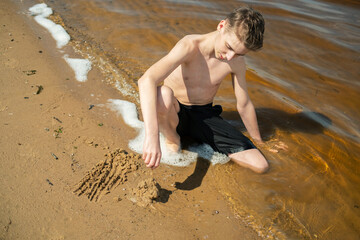 Young boy sits on the sandy lakeside, focused and creative, building a sandcastle under a clear blue sky surrounded by lush green trees, enjoying a warm, sunny day.