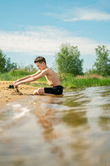 Young boy creating a sandcastle on the shore with focused expression. Warm day at the beach with bright sky and lush greenery in the background, enjoying playful outdoor activity.
