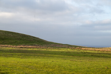 A vibrant green meadow stretches across rolling rural hills under a bright blue summer sky filled with fluffy white clouds, offering a panoramic nature view of the countryside valley