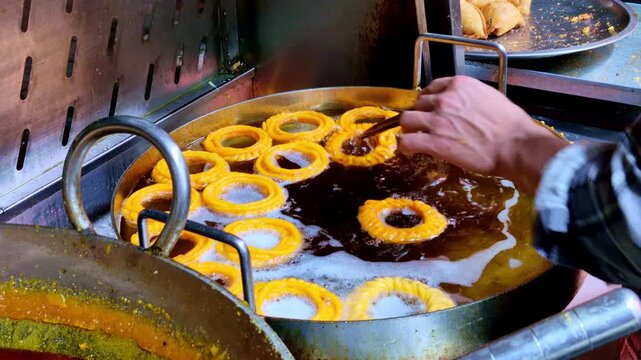 A close-up of a street food vendor frying jalebis imarti in hot oil, showing golden spiral shapes bubbling in a large pan as they are turned with metal tongs.