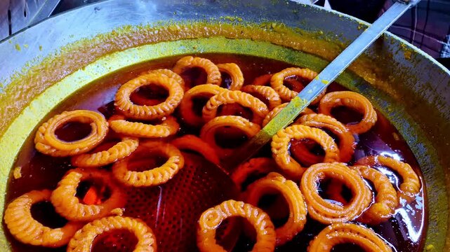A close-up of jalebis frying in a wide pan of hot oil, showing crisp golden rings submerged in bubbling syrupy liquid as they cook evenly.