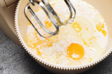Mixing pancake batter closeup. Eggs and milk in bowl with electric mixer beaters. Preparing homemade crepes dough in kitchen. Baking ingredients being blended. Cooking process step at home.