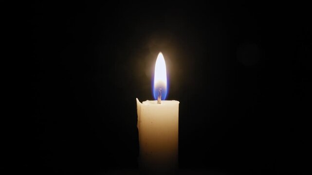 Single white candle burns with a gently flickering flame in the center of the frame. The isolated flame is the only source of light against a completely black background.