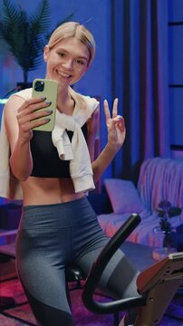 Portrait of charming cheerful athletic fit slender 25-aged blonde in training clothes which photographing herself on smartphone while turning the pedals on bike trainer in cozy night lighted room