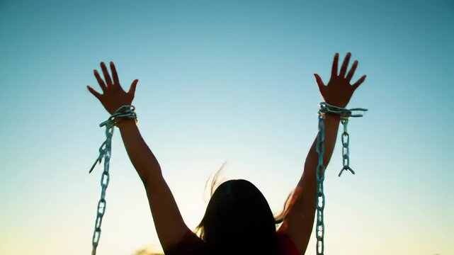 Woman in silhouette with arms raised, breaking metal chains around her wrists against a clear sky, symbolizing liberation and freedom journey