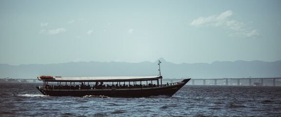 Backlit passenger boat silhouette crossing calm water, crowded deck under roof, distant bridge and hazy mountain range on horizon, minimal seascape with copy space 