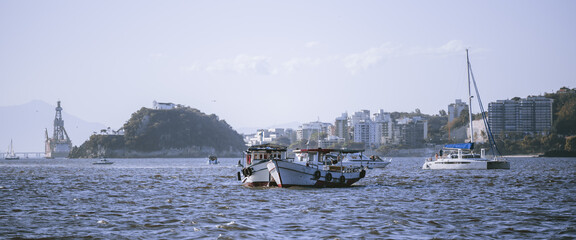 Wide coastal bay with small fishing boat and yacht on rippled water, island hill and city skyline in background, hazy mountains and pale sky, travel and harbor scene