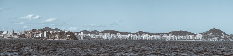Telephoto panoramic view of Niteroi waterfront skyline across Guanabara Bay, modern apartment towers and green hills under blue sky, textured sea foreground, copy space