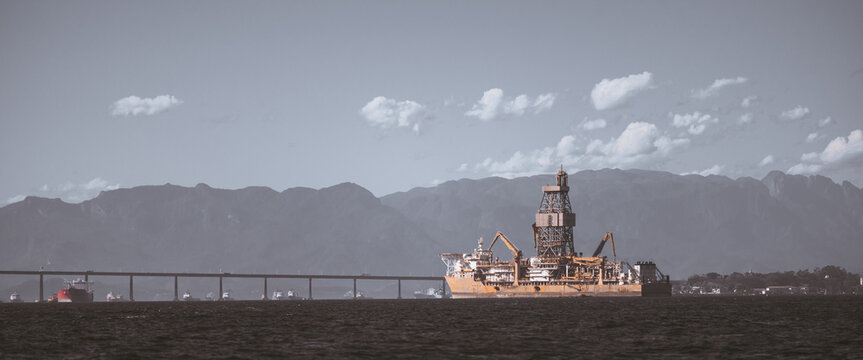 Telephoto view of offshore oil drilling platform ship near a long bridge with hazy mountain backdrop, calm sea, industrial energy concept, wide copy space on the left