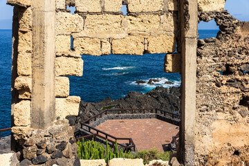 Ruined stone building frame with arched openings overlooking ocean waves, black rocks and hexagonal viewing platform at Costa El Sauzal, Tenerife, Canary Islands, Spain.