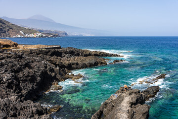 Fototapeta premium Person walking along rugged black volcanic rock coastline with turquoise ocean waves and distant Mount Teide volcano horizon at Costa El Sauzal, Tenerife, Canary Islands, Spain.