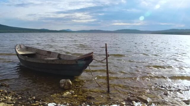 A serene landscape view of the high-mountain Lake Zyuratkul in the Southern Urals, Russia. An old, empty wooden fishing boat is tethered to a post near the rocky shore, with calm water and forested mo