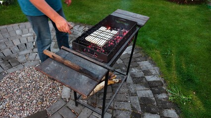 On a tiled area near a grassy lawn there is a metal brazier in which coals are smoldering and the light burns. A man flips a grate of cheese in a pita bread. Autumn evening twilight