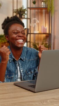 African American young woman celebrates good results from an online test and victory in a digital laptop game while sitting at table on home sofa. Black girl joyfully reacts to success, uplifting news