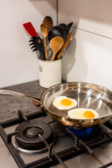 A stainless steel pan frying two eggs with kitchen utensils in the background
