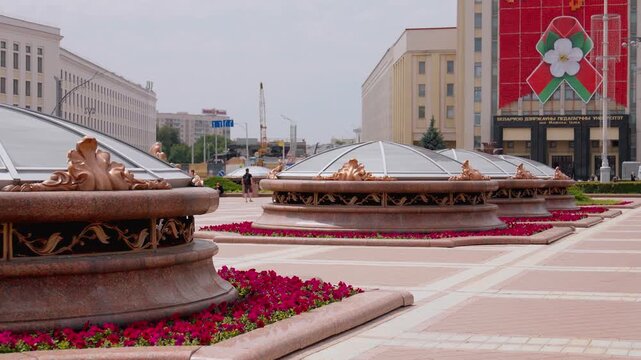 Urban landscape featuring the unique architecture of independence square in minsk, belarus, with decorative fountains, blooming red flowers, and surrounding government buildings on a sunny day