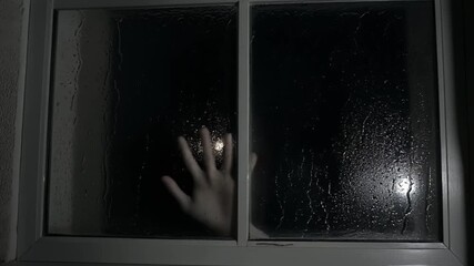 A hand pressed against a rain-streaked window at night, with a faint light source creating a mysterious glow.