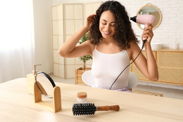 Beautiful young African-American woman using modern hair dryer at table in bathroom