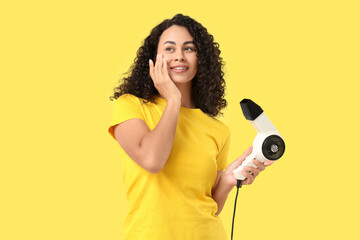 Beautiful young African-American woman with modern hair dryer on yellow background