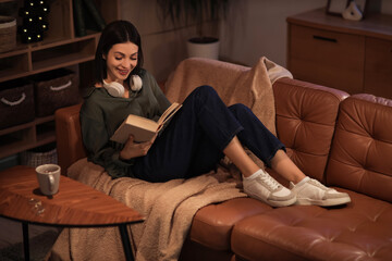 Obraz premium Beautiful young woman with headphones reading book and sitting on sofa in living room at night