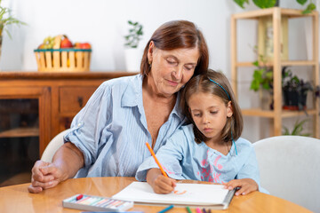 Grandmother watching girl drawing with colored pencil at home showing encouragement and admiration while child focuses on creative activity