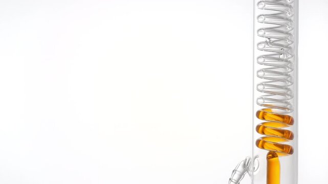 Orange fluid flows through a coil lab condenser, filling the coiled glass tube on the right side, leaving the left side as copy space on white surface | Marigold infusion cosmetic science concept