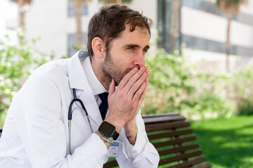 Healthcare professional experiencing stress and burnout, male doctor with hands clasped, expressing worry and exhaustion while sitting alone on a bench outdoors