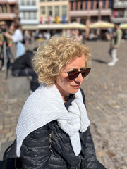 attractive sixty year old mature woman with curly blonde hair sitting at a bench and enjoying the sun, wearing sunglasses