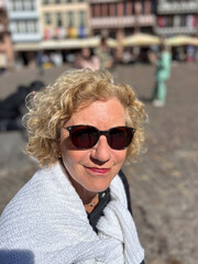 attractive sixty year old mature woman with curly blonde hair sitting at a bench and enjoying the sun, wearing sunglasses