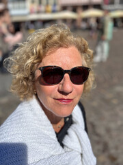 attractive sixty year old mature woman with curly blonde hair sitting at a bench and enjoying the sun, wearing sunglasses
