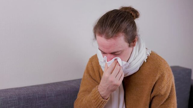 Sick man with a red nose blowing his nose into a tissue while sitting on the sofa at home having the flu. High quality 4k footage