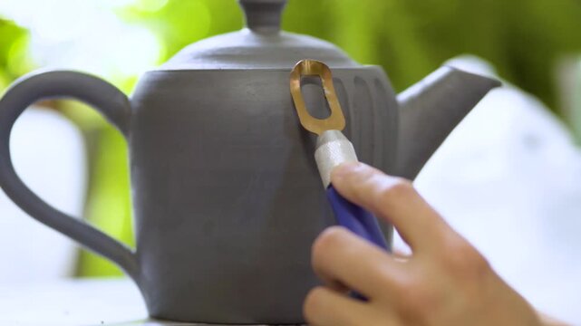 Skilled craftswoman meticulously carving decorative patterns on wheel-thrown clay teapot. Meditative pottery work in sunlit studio.