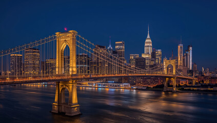 Fototapeta premium Golden illuminated suspension bridge spanning calm river with illuminated skyline at night