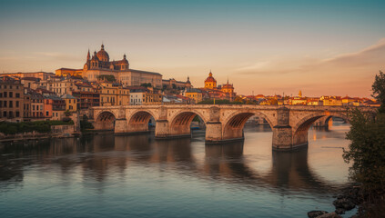 Fototapeta premium Historic stone bridge over calm river with warm sunset light and city skyline