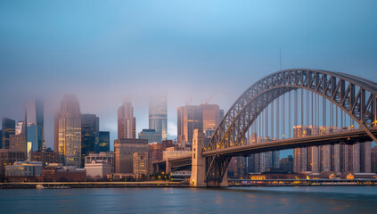 Obraz premium City skyline at misty dawn with arched steel bridge and soft warm light