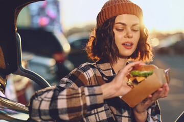 Woman in beanie holding burger by car trunk at sunset, candid urban portrait with authenticity and...