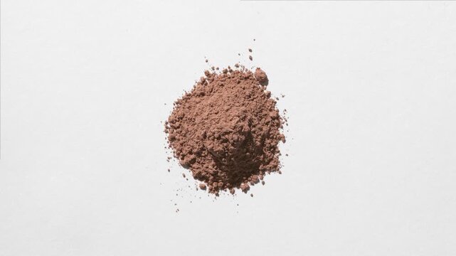 Overhead frame of a pile of brown powder on a dim grey background lit with dynamic orbiting light that casts shifting shadows | Carob Powder Commercial Concept