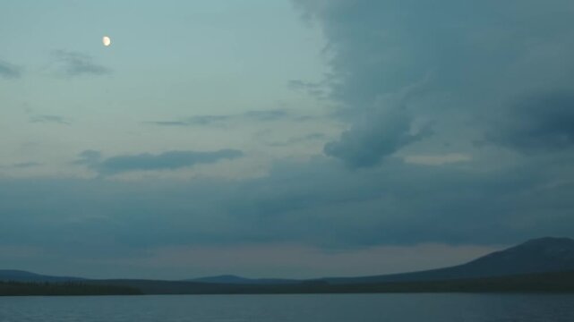A wide-angle shot of a tranquil lake with rolling hills in the distance under a cloudy, twilight sky. A crescent moon is visible in the upper left corner. The serene landscape captures the natural bea