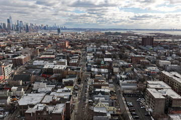 Aerial drone photo from Jersey City featuring Manhattan skyline views, dense city blocks, waterfront buildings, and modern New York City architecture under dynamic daytime cloud cover.                