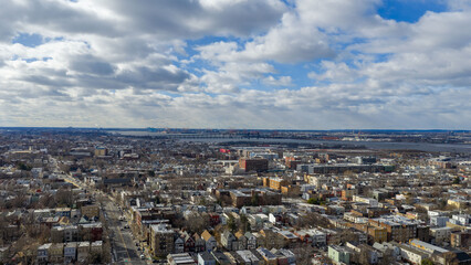 Aerial drone photo from Jersey City featuring Manhattan skyline views, dense city blocks, waterfront buildings, and modern New York City architecture under dynamic daytime cloud cover.                
