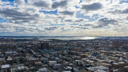 Aerial drone photo from Jersey City featuring Manhattan skyline views, dense city blocks, waterfront buildings, and modern New York City architecture under dynamic daytime cloud cover.                