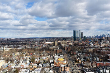 Aerial drone photo from Jersey City featuring Manhattan skyline views, dense city blocks, waterfront buildings, and modern New York City architecture under dynamic daytime cloud cover.                