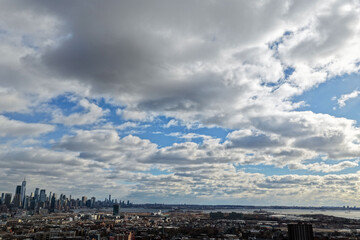 Aerial drone photo from Jersey City featuring Manhattan skyline views, dense city blocks, waterfront buildings, and modern New York City architecture under dynamic daytime cloud cover.                