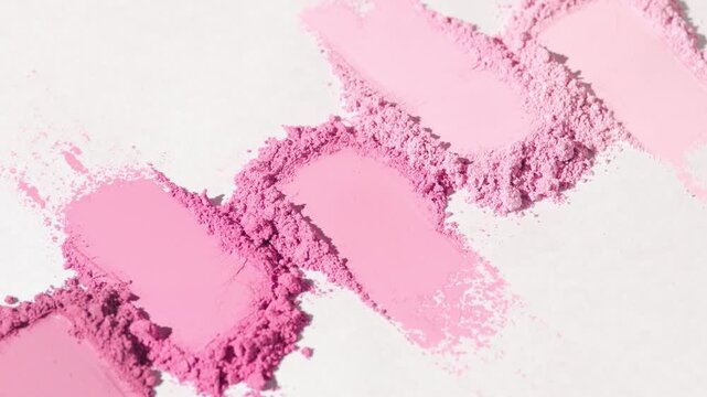 Vertical orbit macro close-up of five pale to pink loose powder smears organized in line on a light gray background illuminated by rotating light forming dynamic shadows