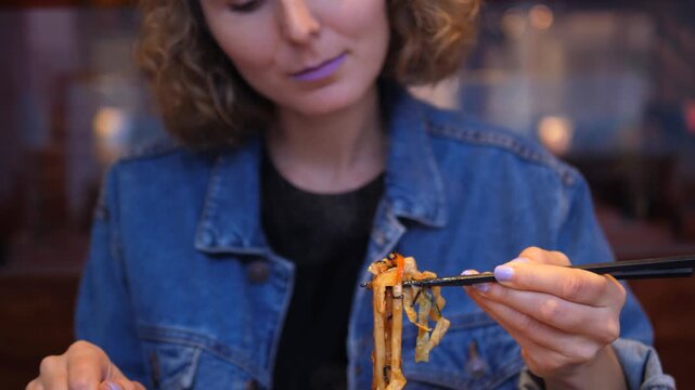Young woman in a denim jacket enjoying a delicious meal of asian noodles and vegetables with chopsticks, with a plate of sushi rolls on the table in a traditional restaurant