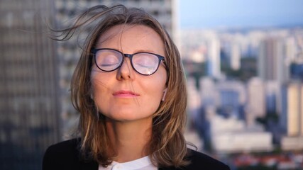 Young businesswoman with closed eyes and hair blowing in the wind listening to music on wireless earbuds, meditating and relaxing on a skyscraper rooftop with a beautiful city view at sunset